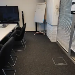 Wnętrze biura z białym stołem konferencyjnym, czarnymi krzesłami, tablicą suchościeralną z prostymi rysunkami i zasłoniętym oknem żaluzjami.