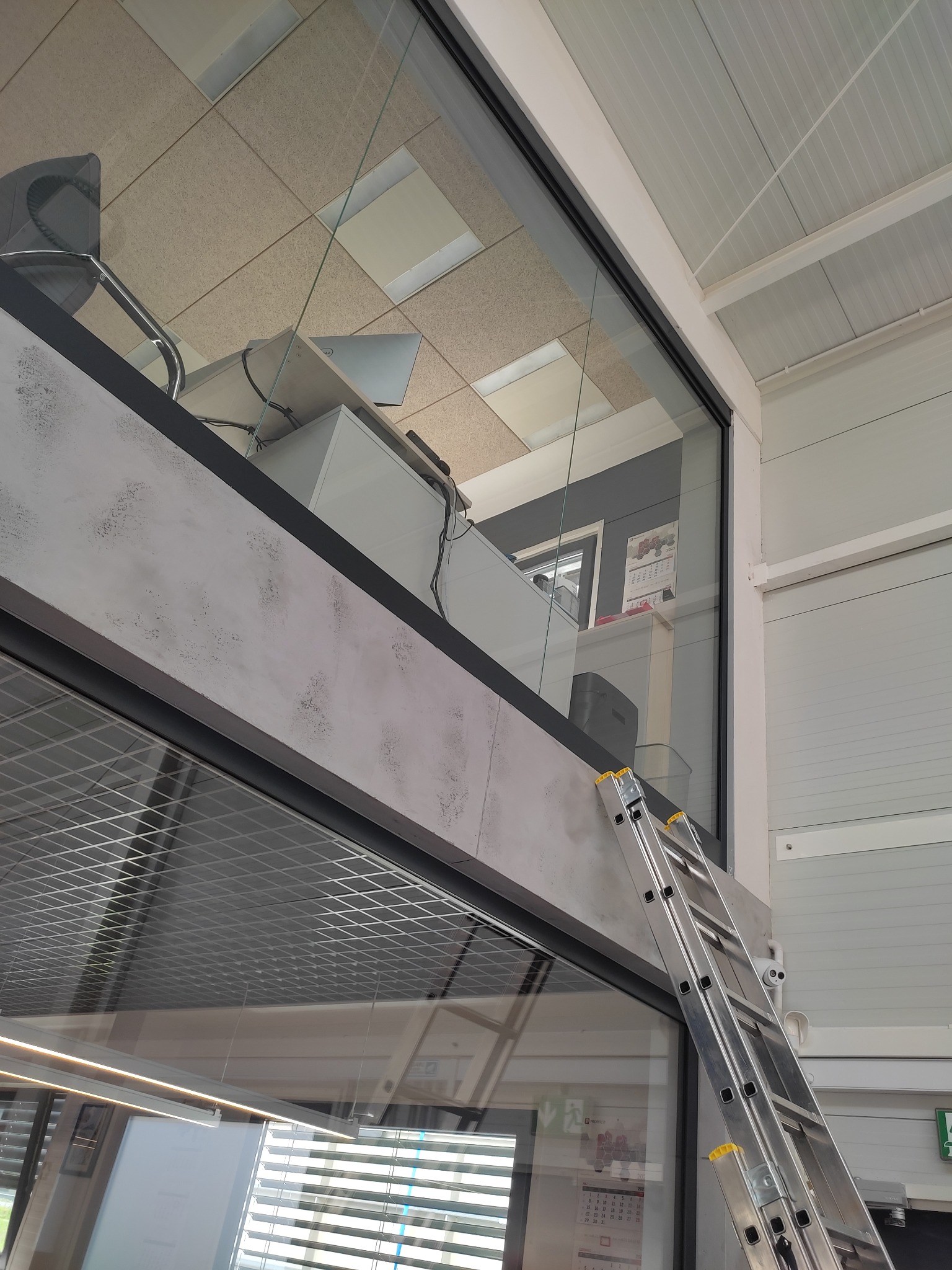 Aluminiowa drabina oparta o przeszkloną ścianę biura na antresoli, widok z dołu podczas prac na wysokości.