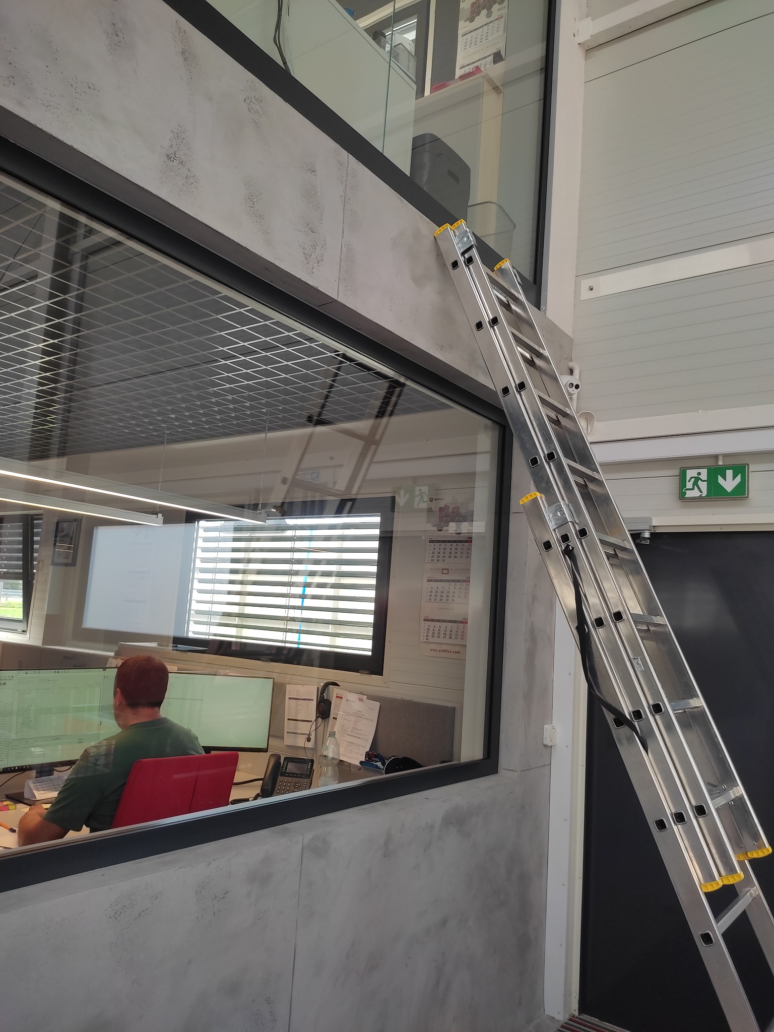 Aluminiowa drabina oparta o ścianę z oknem w biurze; widoczny pracownik przy biurku,  sufit z kratką, lampy liniowe i znak ewakuacyjny.