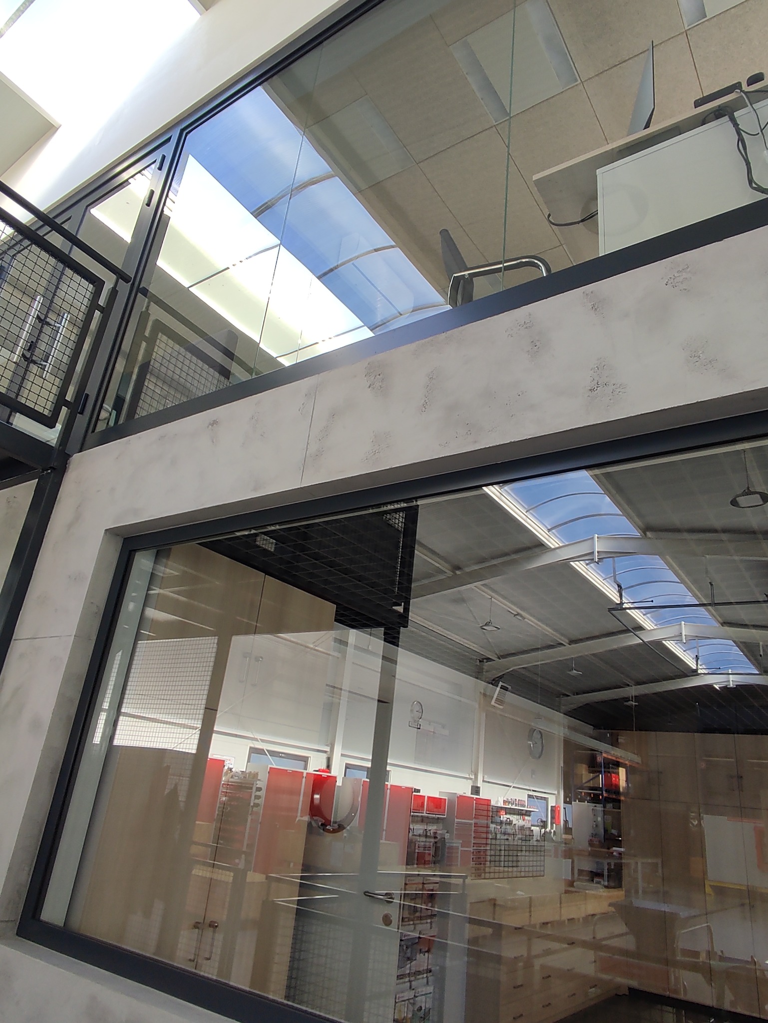 Wnętrze nowoczesnego biura w Warszawie widziane przez duże okno z czarną ramą. Widoczne biurka, krzesła i regały z produktami. Przez okna w dachu wpada naturalne światło.