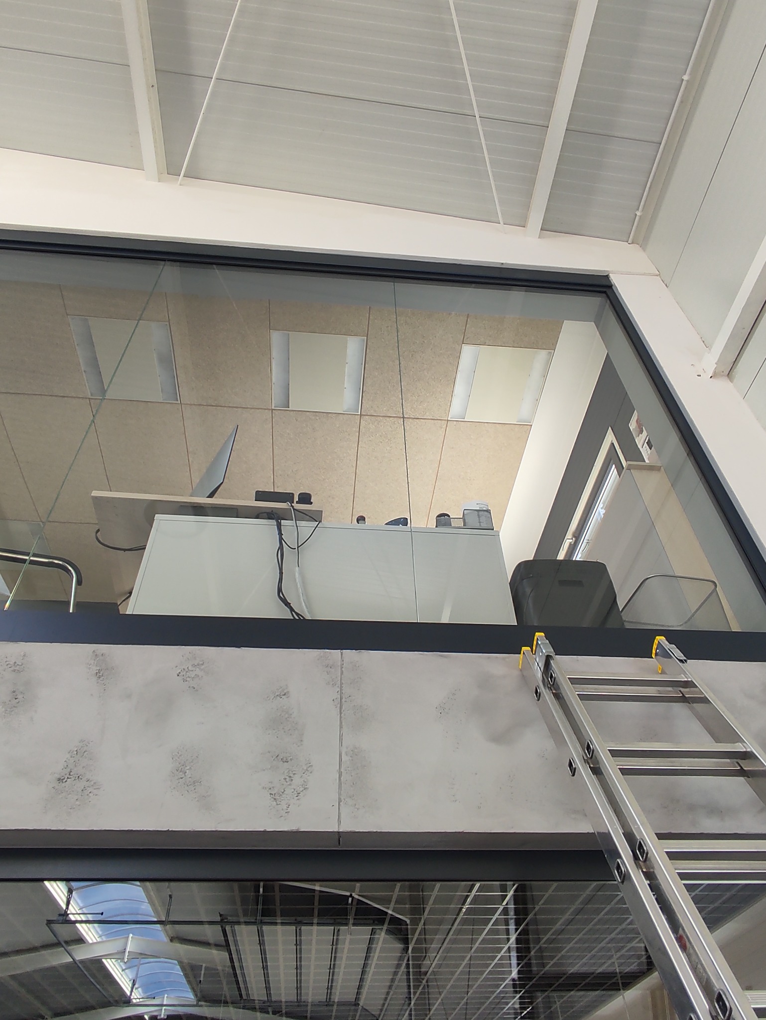 Wnętrze biura widziane z dołu przez szklaną ścianę, z widoczną drabiną opartą o ścianę, sugerującą prace porządkowe lub konserwacyjne.