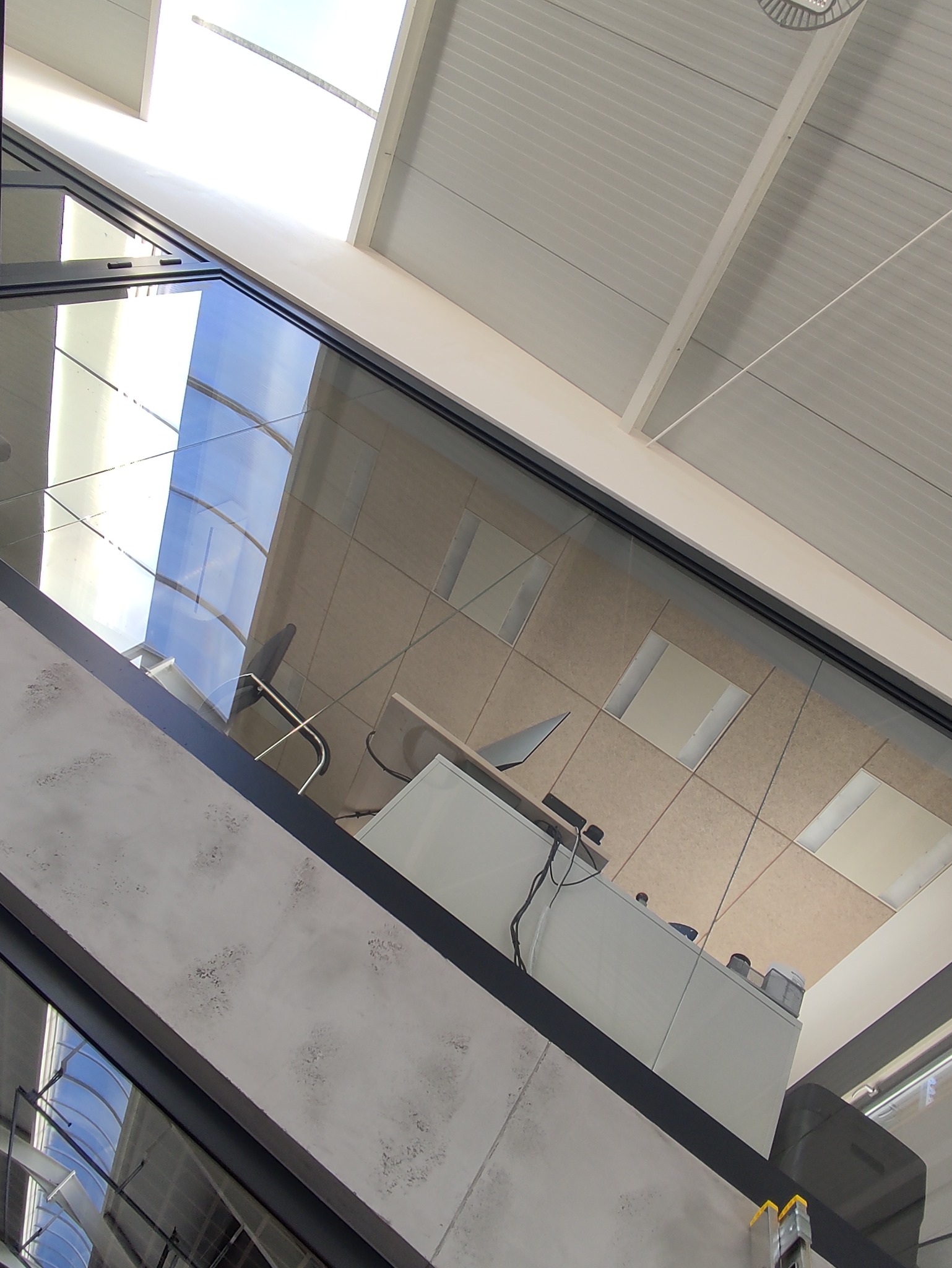 Wnętrze biura z widokiem na sufit z oknami dachowymi, biurko z komputerem i krzesłem, widziane z perspektywy nisko położonej ściany z elementami szklanymi.