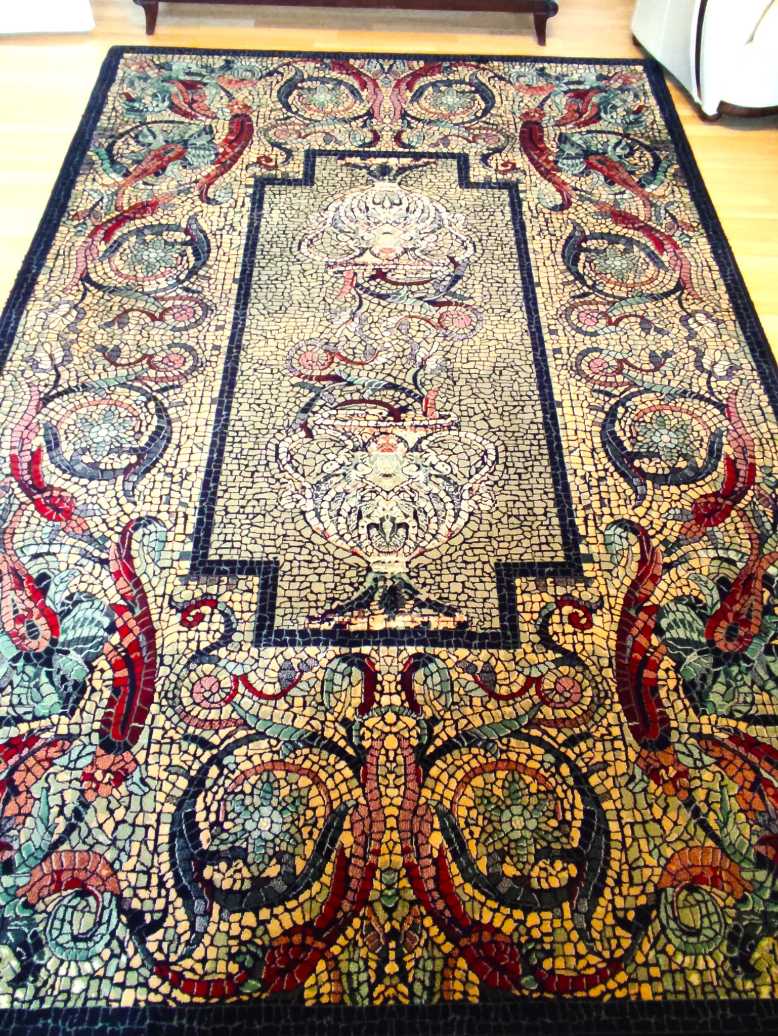 Mozaikowy dywan z motywami roślinnymi i wazonem w centralnej części, ułożony na drewnianej podłodze.