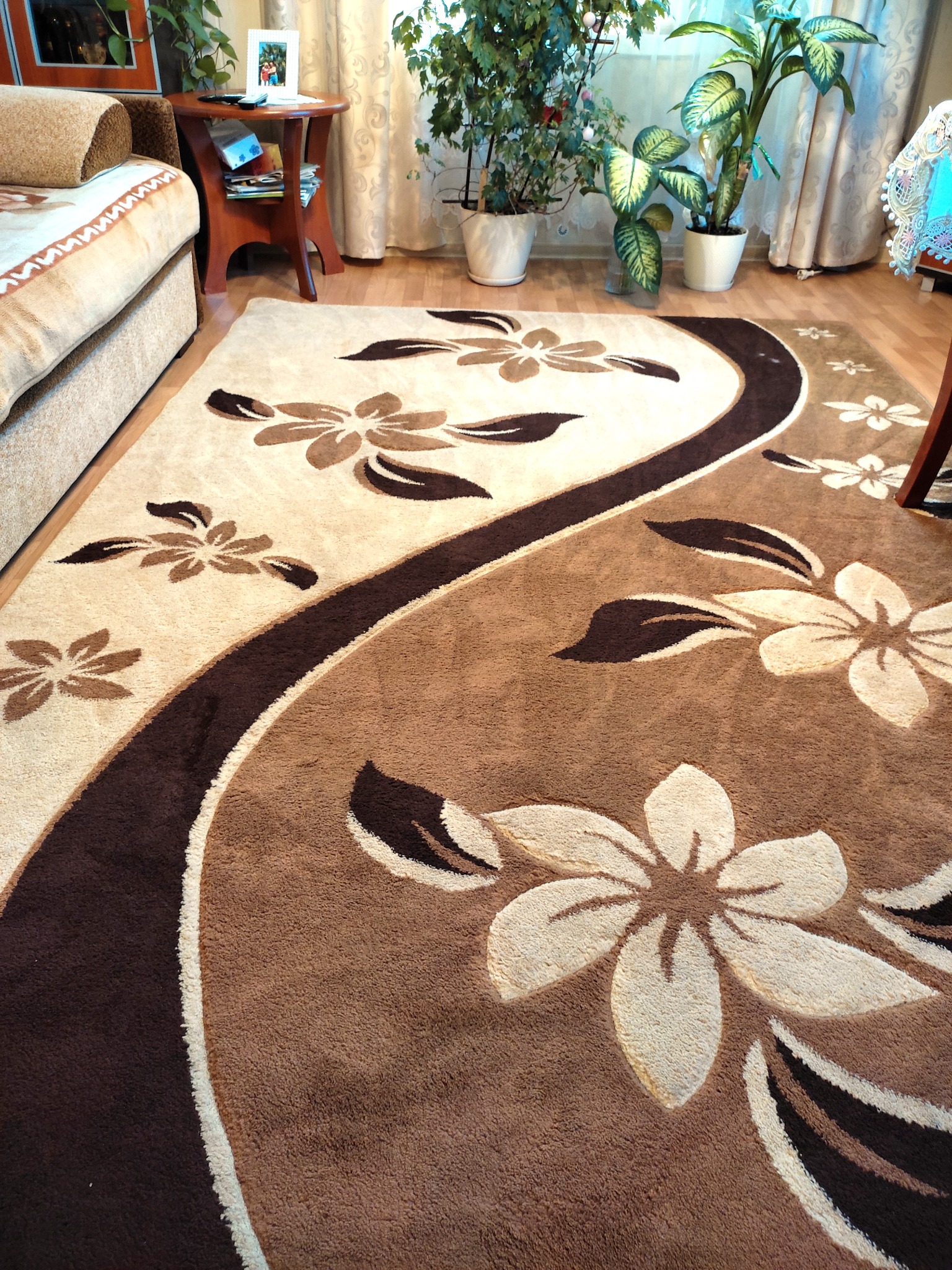 Beżowo-brązowy dywan z motywem kwiatowym, leżący na parkiecie w jasnym wnętrzu z roślinami doniczkowymi i meblami.
