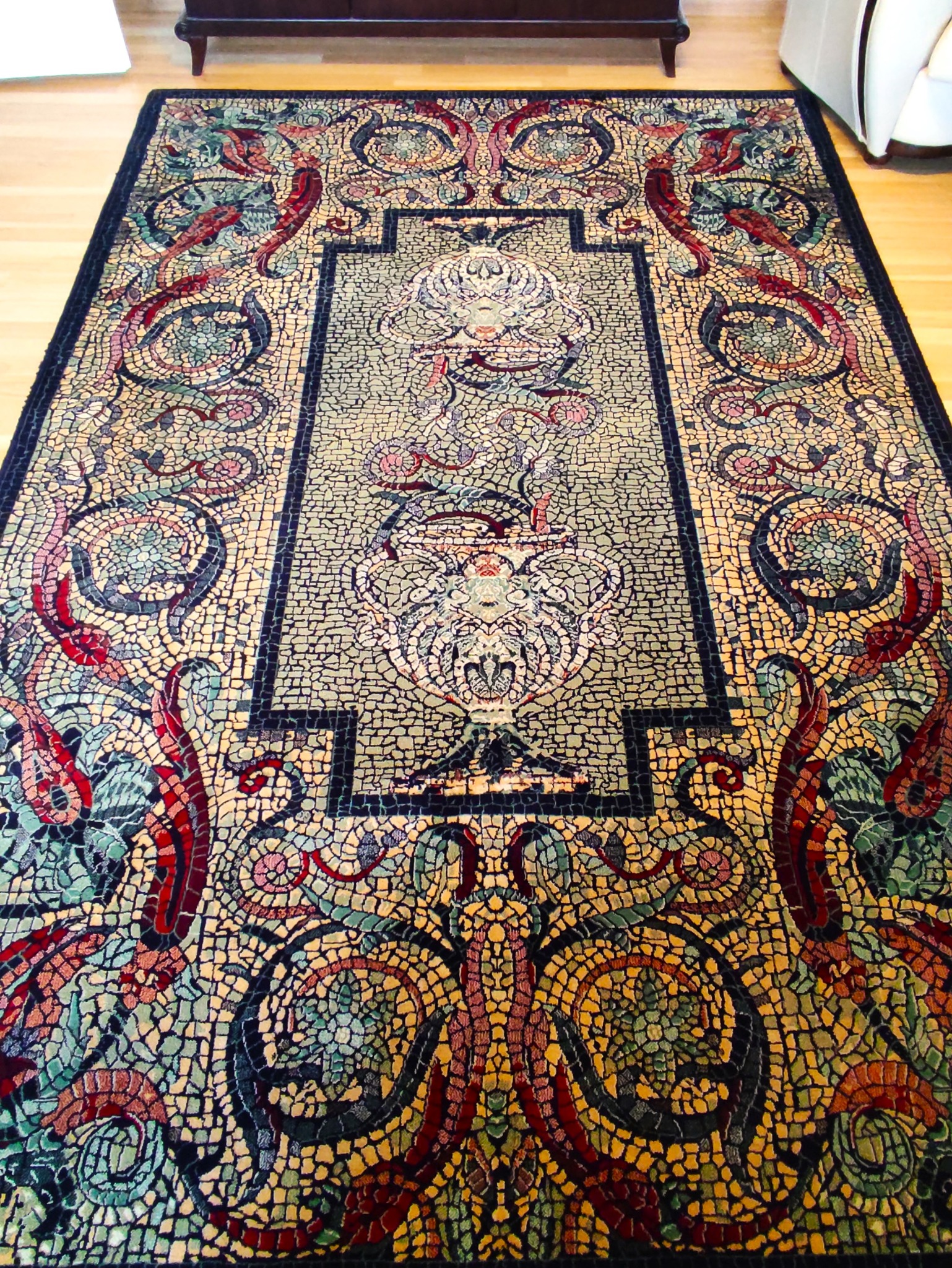 Prostokątny dywan z misternym wzorem mozaikowym w odcieniach beżu, zieleni, czerwieni i czerni, leżący na drewnianej podłodze, z fragmentem białego mebla w tle.