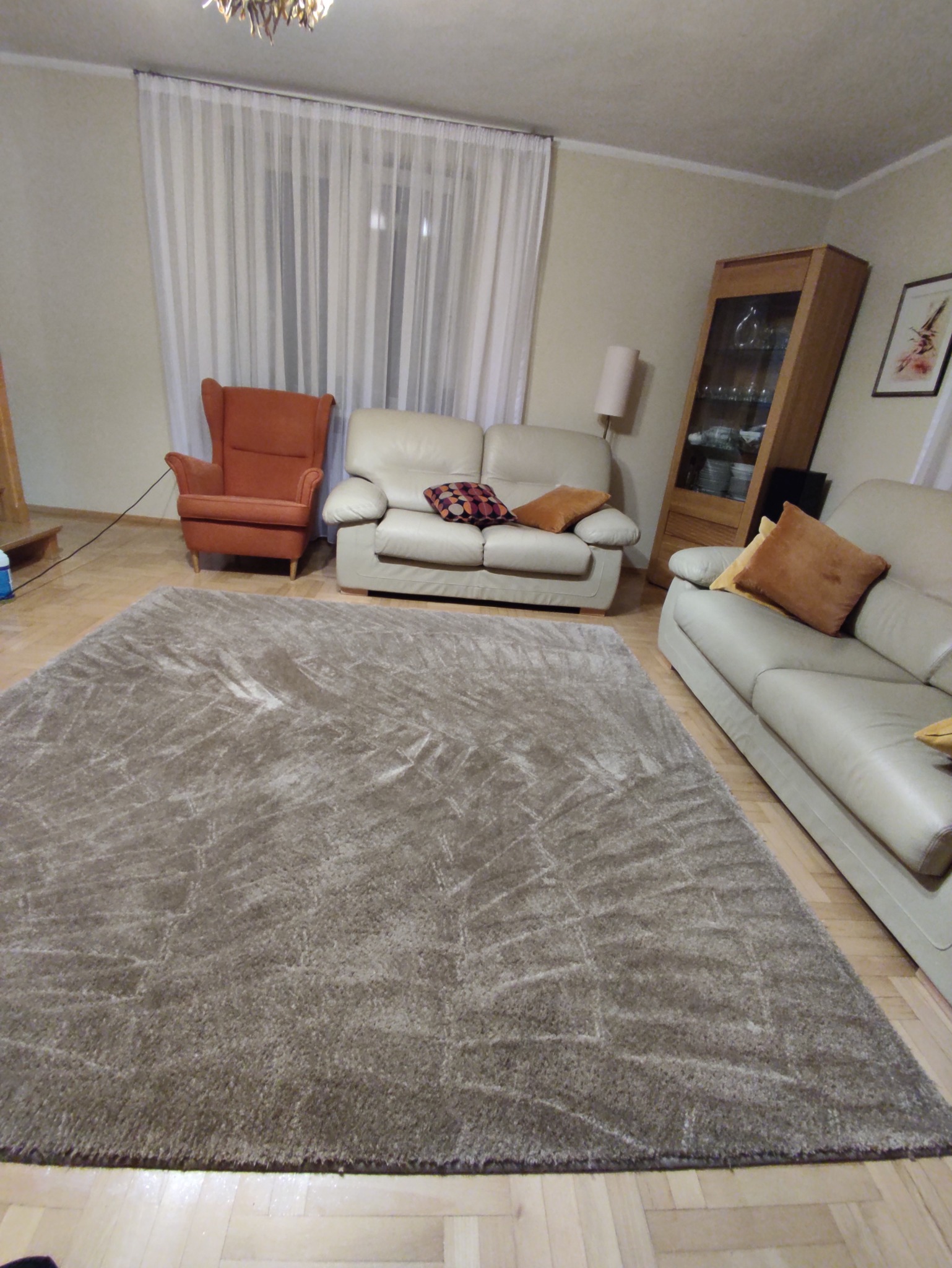 Beżowy dywan shaggy z geometrycznym wzorem na parkiecie, w tle kremowe sofy, fotel i witryna z oświetleniem.