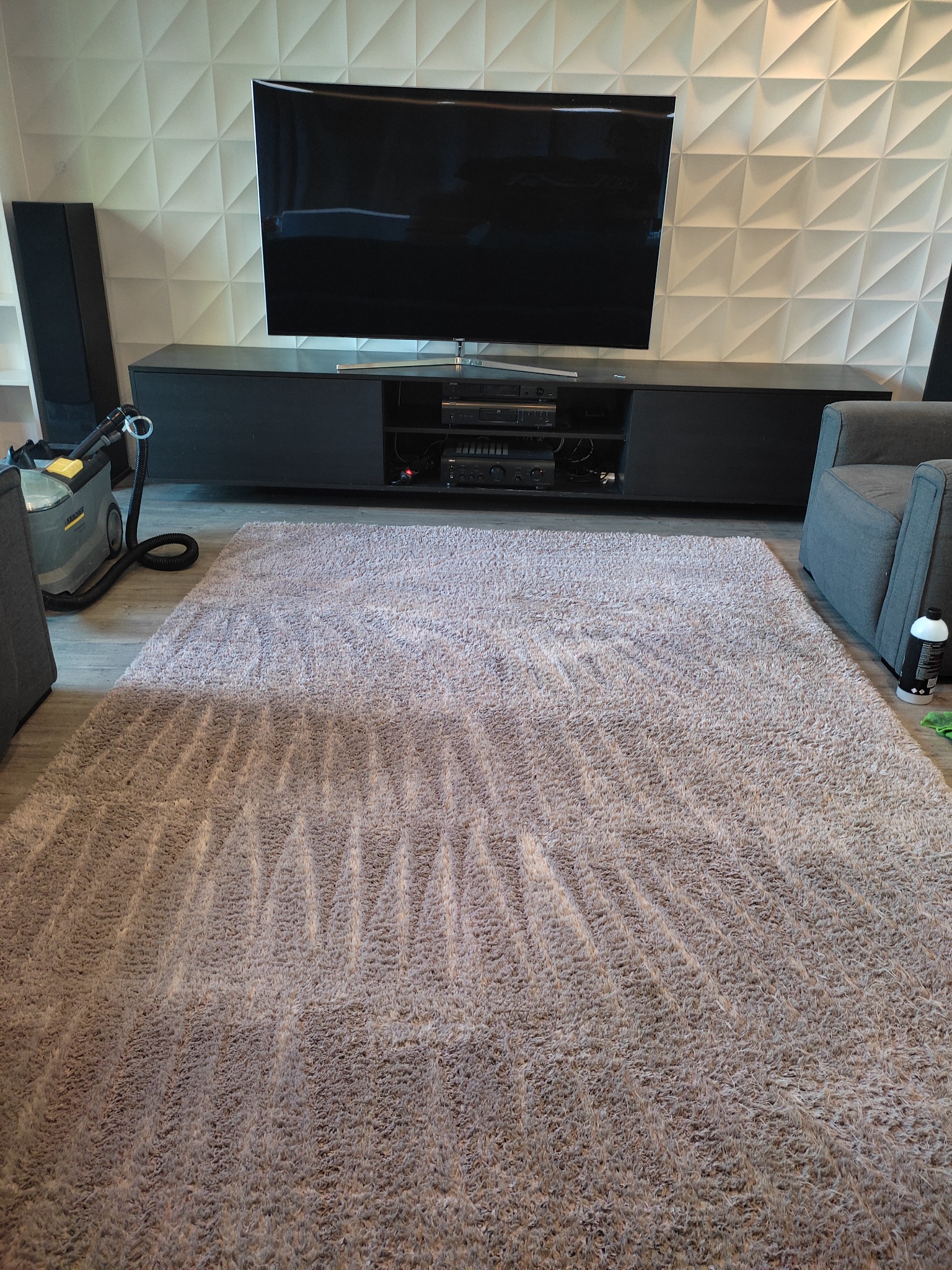 Świeżo wyprany, beżowy dywan z widocznymi śladami czyszczenia, obok szary fotel i odkurzacz piorący Karcher, na tle ściany z geometrycznym wzorem 3D.