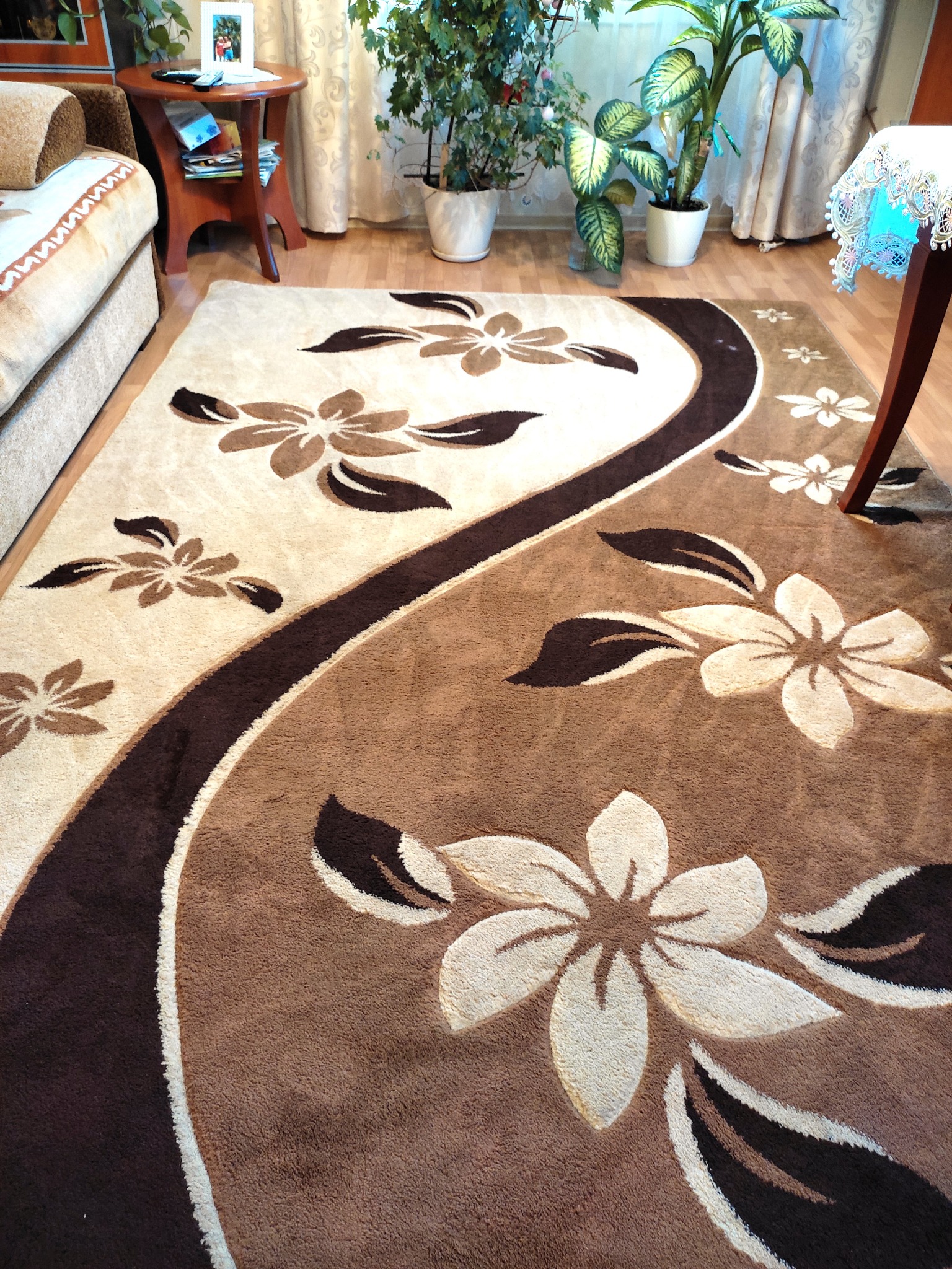 Beżowo-brązowy dywan z motywem kwiatowym, ułożony na drewnianej podłodze w salonie, widoczny fragment sofy i roślin doniczkowych.