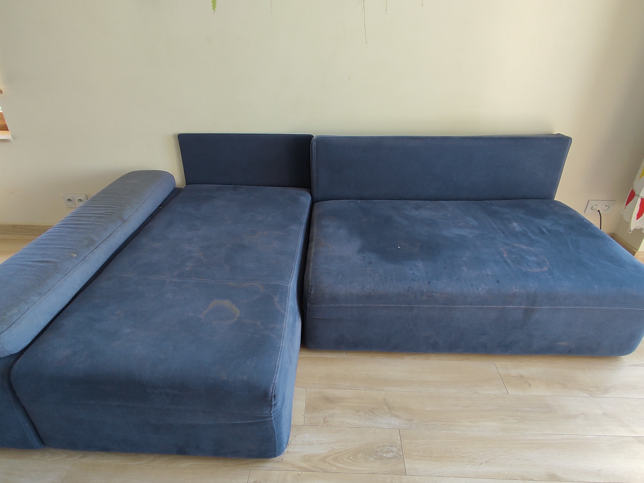 Niebieska, modułowa kanapa z widocznymi plamami na tapicerce, stojąca na panelach podłogowych, sfotografowana w słabym oświetleniu.