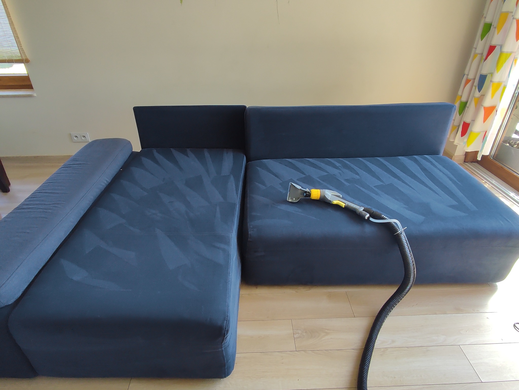 Granatowa, modułowa kanapa narożna z widocznymi śladami czyszczenia na powierzchni, obok leży dysza odkurzacza piorącego z wężem. W tle okno z kolorowymi zasłonami.