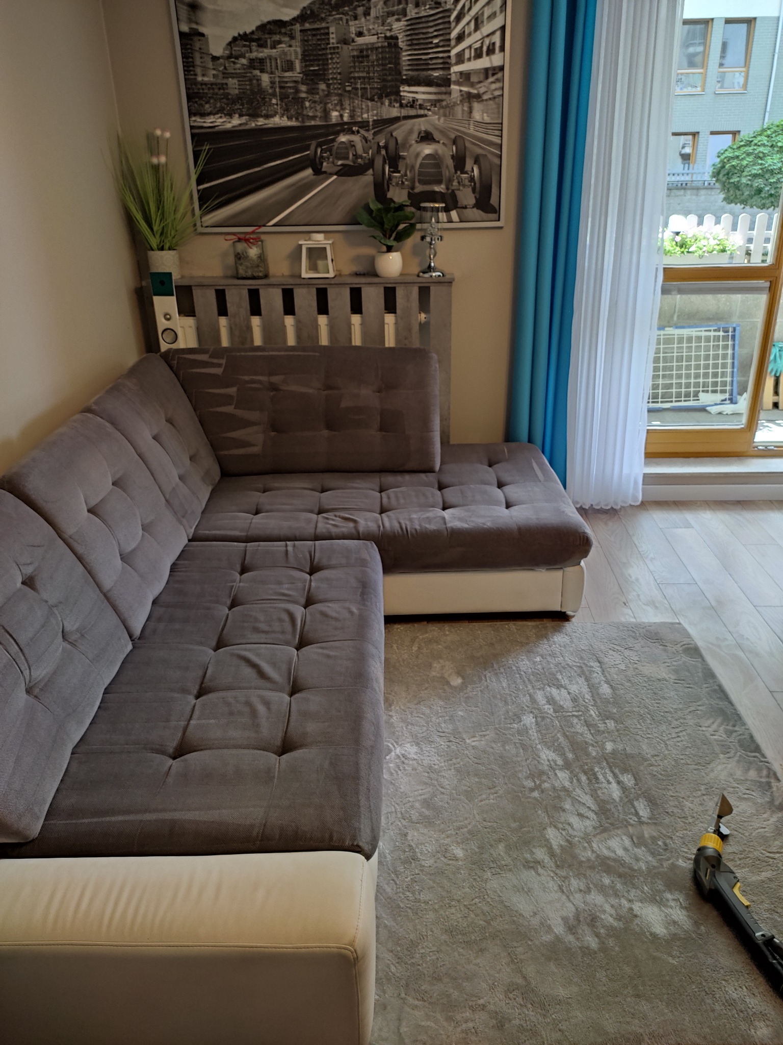 Szara narożna sofa z tapicerowanymi guzikami i jasnym dywanem po czyszczeniu, widoczny fragment odkurzacza piorącego w prawym dolnym rogu, w tle obraz z motywem wyścigowym i okno z błękitnymi...