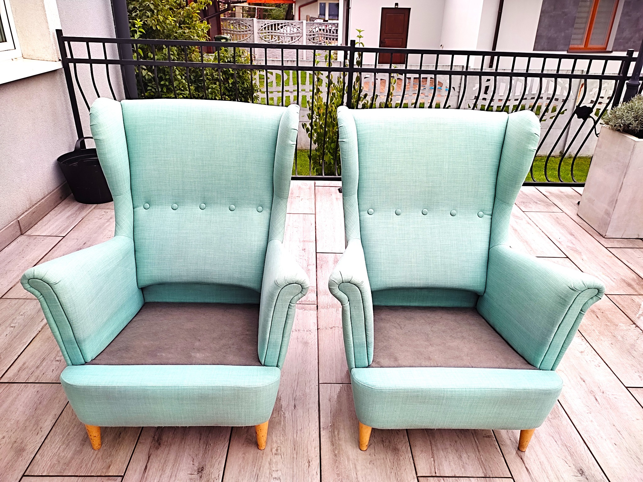Dwa identyczne, miętowe fotele z wysokim oparciem, ustawione symetrycznie na drewnianym tarasie z widokiem na ogrodzenie i budynek mieszkalny.