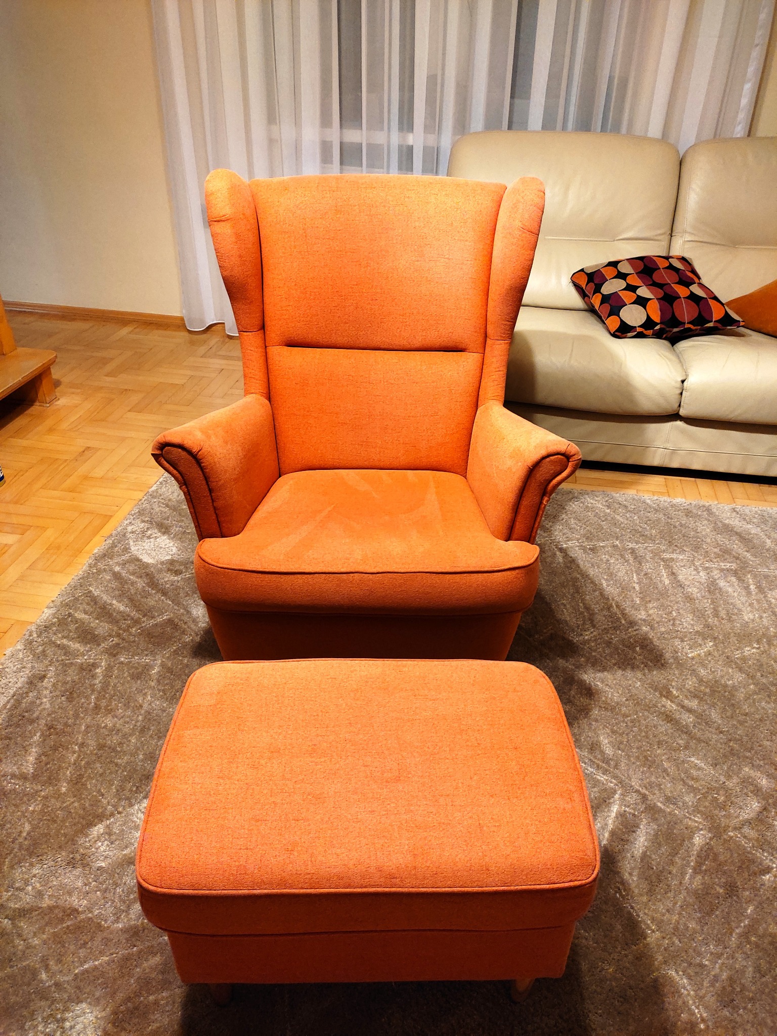 Pomarańczowy fotel uszak z podnóżkiem na beżowym dywanie shaggy, w tle skórzana kanapa i parkiet.
