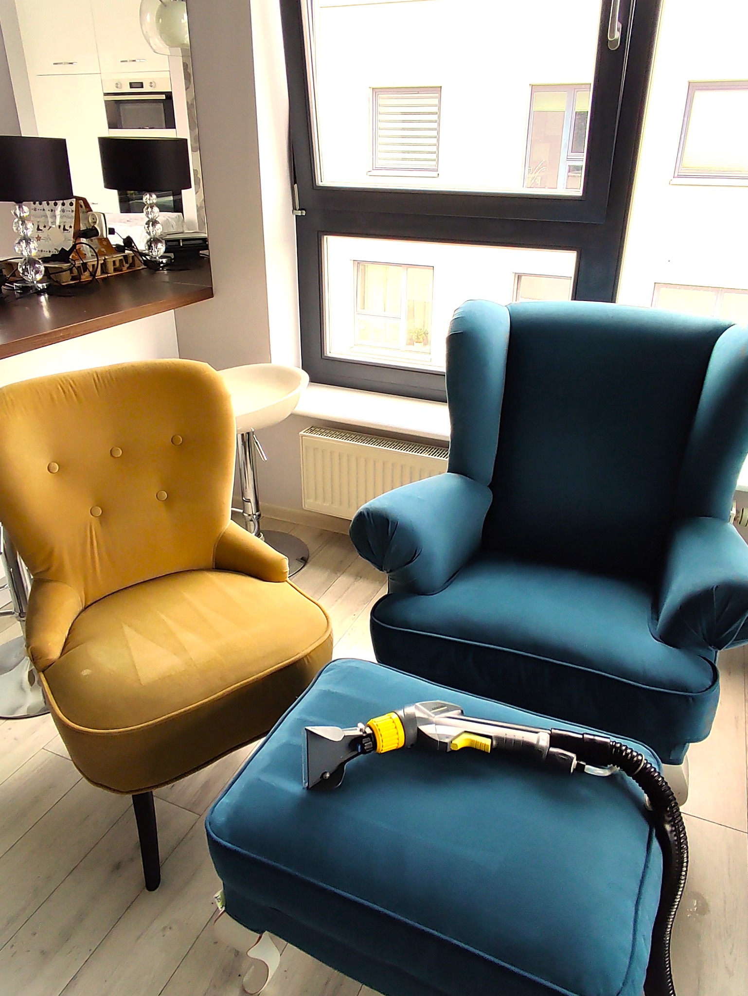 Żółty fotel klubowy i turkusowy fotel uszak obok pufy z dyszą do czyszczenia tapicerki na tle nowoczesnego salonu z oknem.