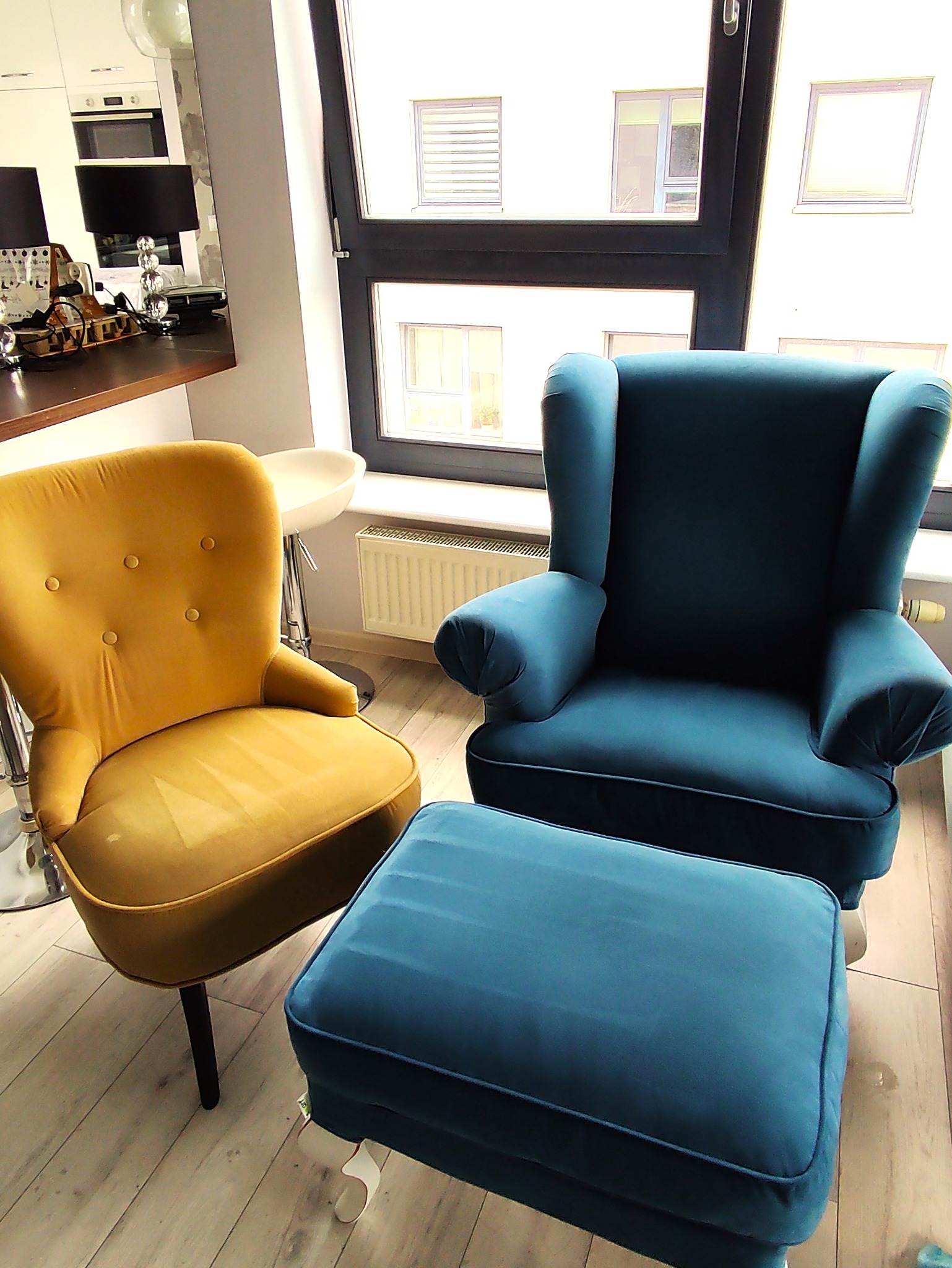 Żółty fotel klubowy i turkusowy fotel uszak z podnóżkiem, ustawione na parkiecie w jasnym wnętrzu mieszkania z widokiem na okno.