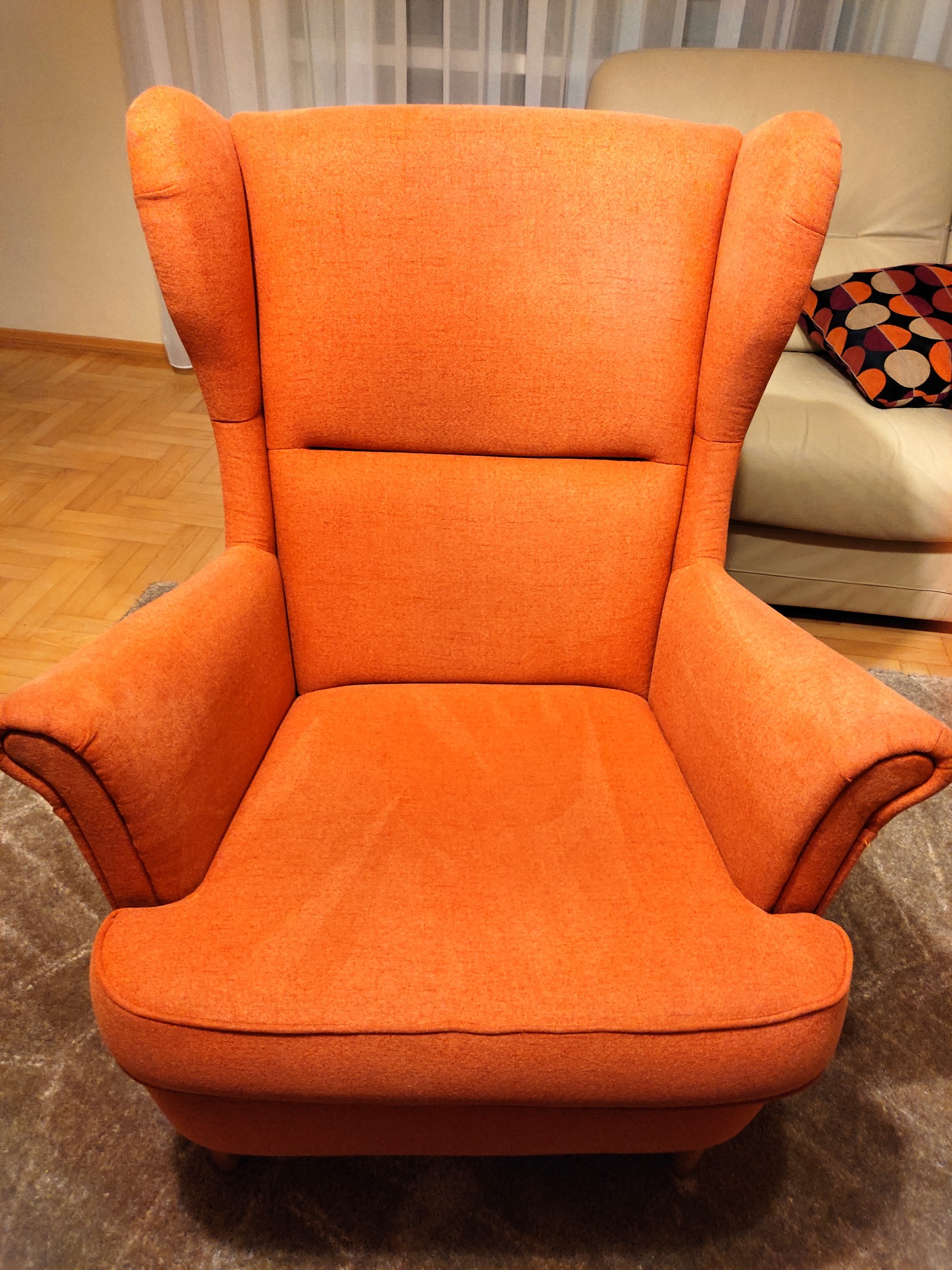 Pomarańczowy fotel typu uszak na tle parkietu i jasnej sofy w salonie w Warszawie. Widoczny fragment dywanu pod fotelem.