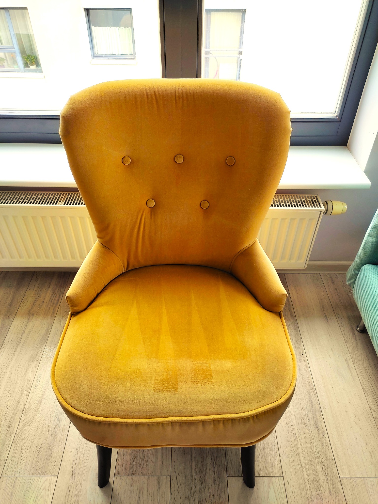 Żółty, welurowy fotel typu uszak na tle okna i grzejnika, widoczne ślady użytkowania na siedzisku.