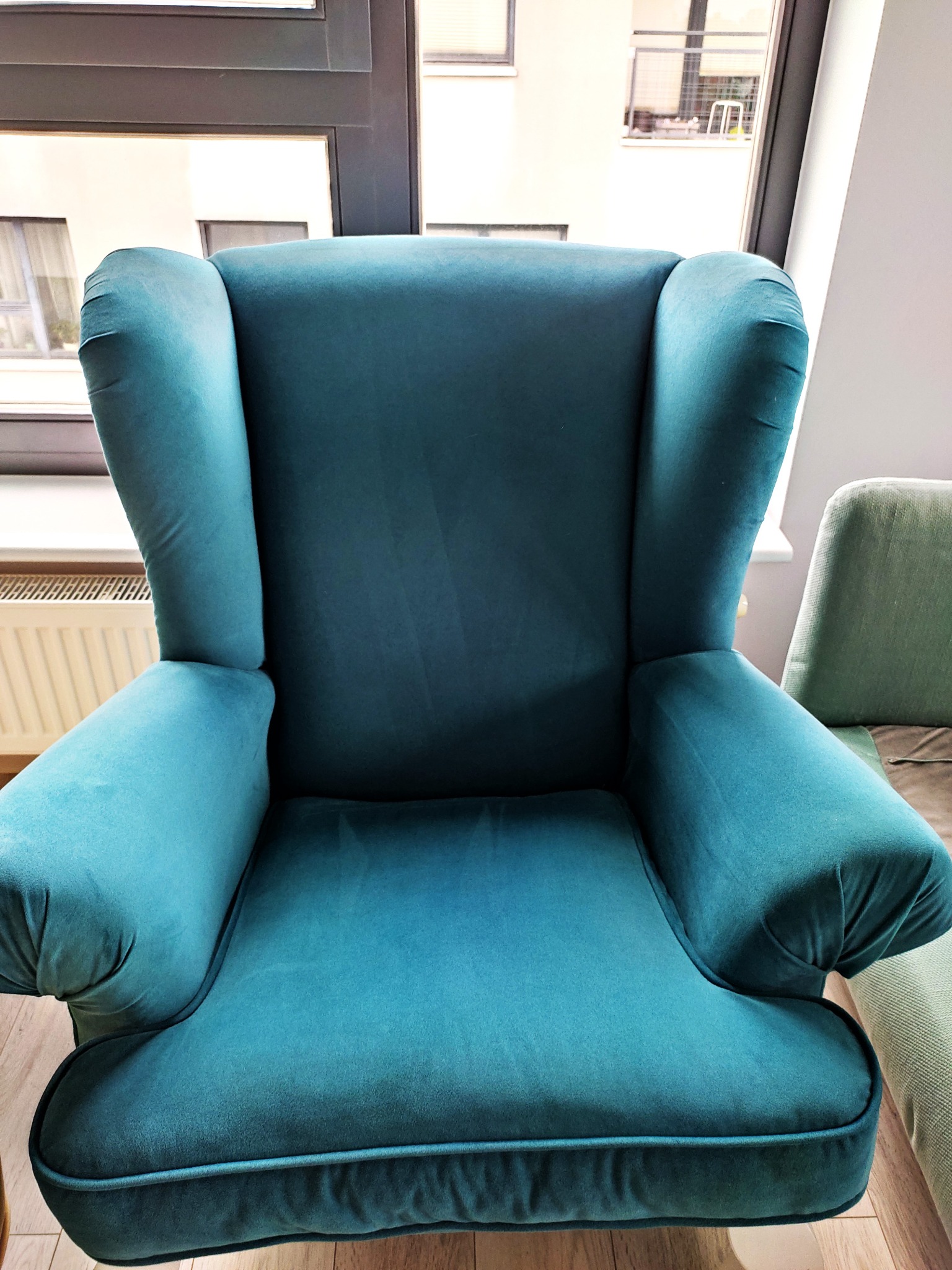 Turkusowy fotel uszak z welurową tapicerką, ustawiony w jasnym wnętrzu, widok z przodu.