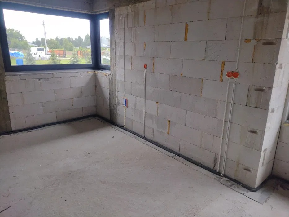 Surowy stan wnętrza z widocznymi przewodami elektrycznymi w ścianie z betonu komórkowego, nieotynkowane ściany, okno z widokiem na zewnątrz.