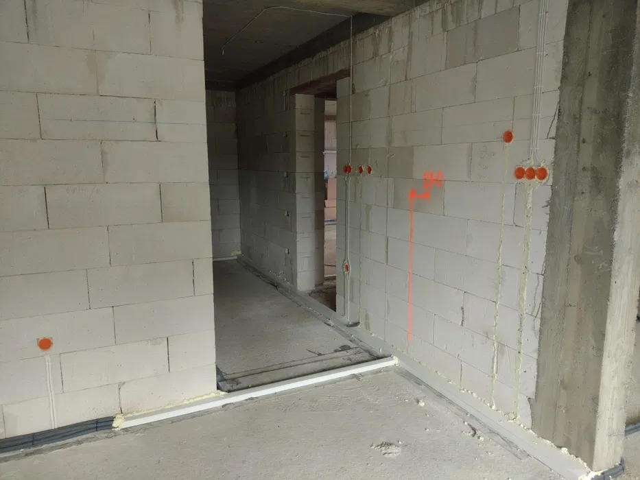 Surowy stan wnętrza budynku w trakcie prac: widoczne kable elektryczne wyprowadzone ze ściany z betonu komórkowego, pomarańczowe oznaczenia na ścianie, listwy przypodłogowe i peszle z kablami...