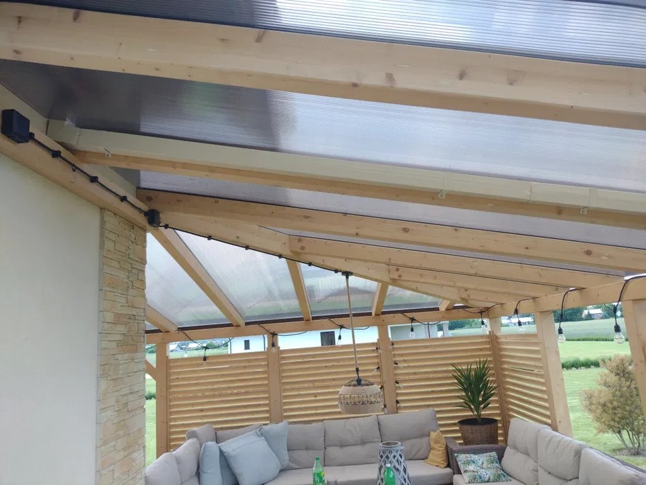 Zadaszony taras z drewnianą konstrukcją, przezroczystym poliwęglanowym dachem, oświetleniem w postaci wiszących żarówek i zewnętrznym gniazdkiem elektrycznym na belce