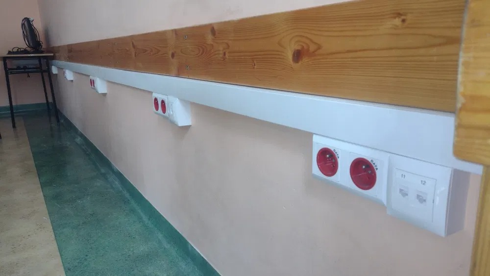 Rząd gniazdek elektrycznych i gniazd sieciowych DATA zamontowanych pod drewnianą listwą przyścienną wzdłuż ściany o jasnoróżowym odcieniu, widoczne w tle biurko z wentylatorem.