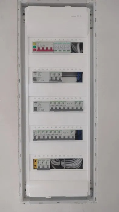 Zamknięta, biała skrzynka rozdzielcza z bezpiecznikami i okablowaniem, zamontowana na białej ścianie, widoczne różne typy zabezpieczeń i oznaczenia numerów obwodów.