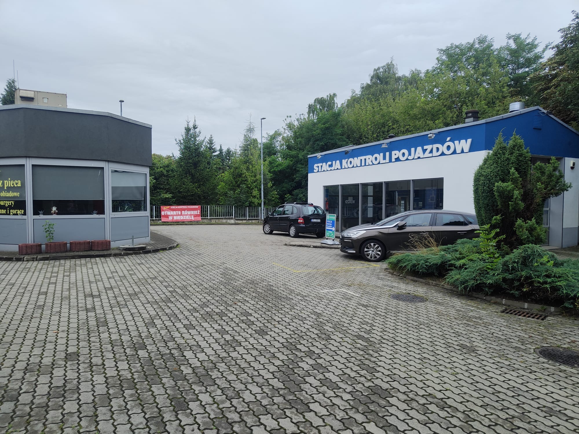Budynek Stacji Kontroli Pojazdów z dwoma samochodami zaparkowanymi na zewnątrz, widok z zewnątrz na szarym brukowanym placu.