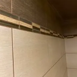 Ściana wyłożona jasnymi, kwadratowymi płytkami ceramicznymi z delikatną strukturą, u góry dekoracyjny pasek z mozaiki w odcieniach brązu i beżu, widoczny fragment sufitu z brązową listwą...