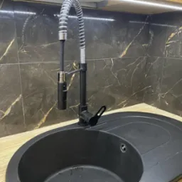 Czarny, okrągły zlew z baterią z wyciąganą wylewką na tle ściany wyłożonej ciemnymi płytkami z marmurkowym wzorem, oświetlonej od góry.