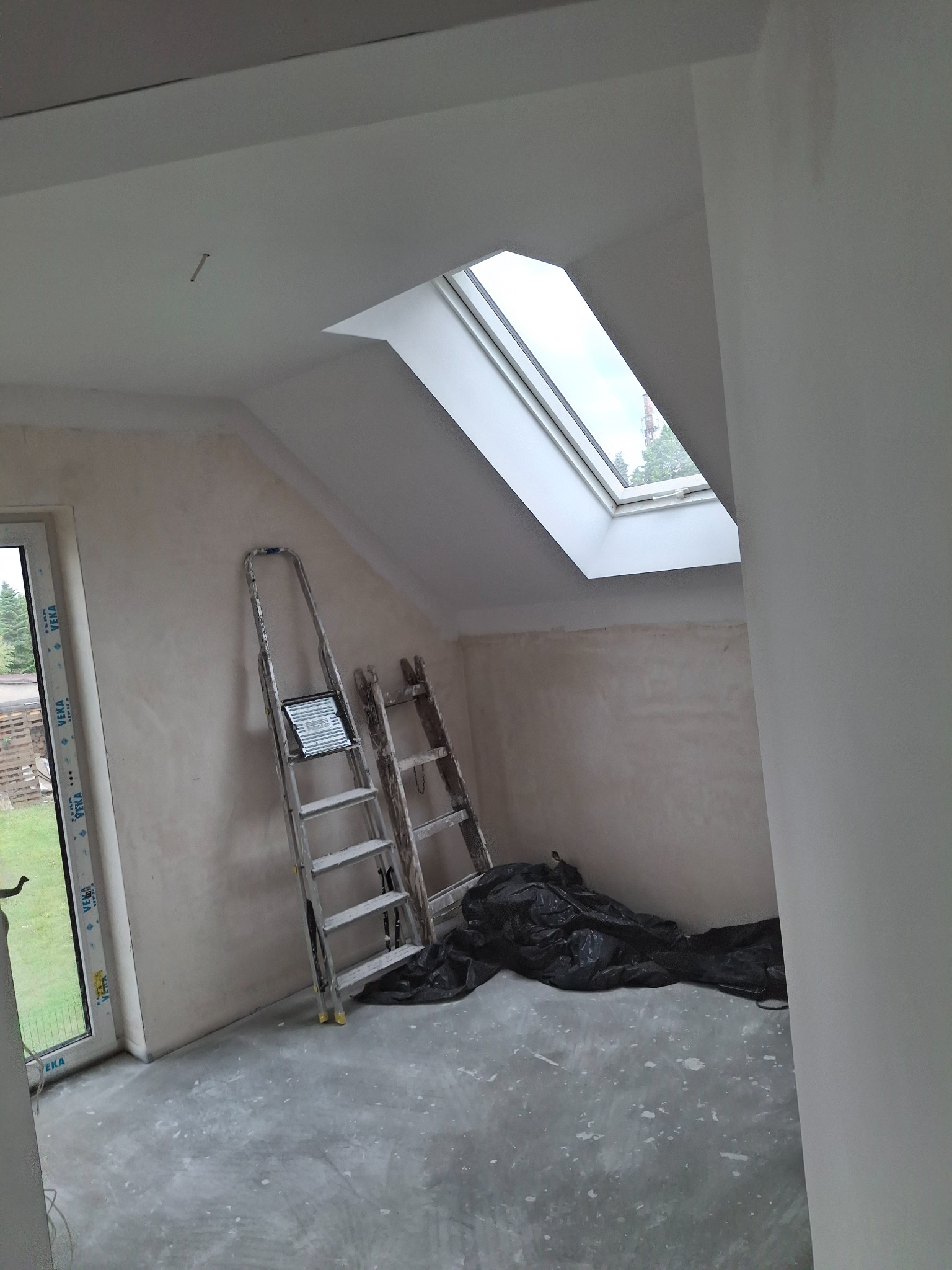 Poddasze ze ścianami po szpachlowaniu, przygotowane do malowania. Dwie drabiny opierają się o ścianę, a przez okno dachowe widać zieleń.