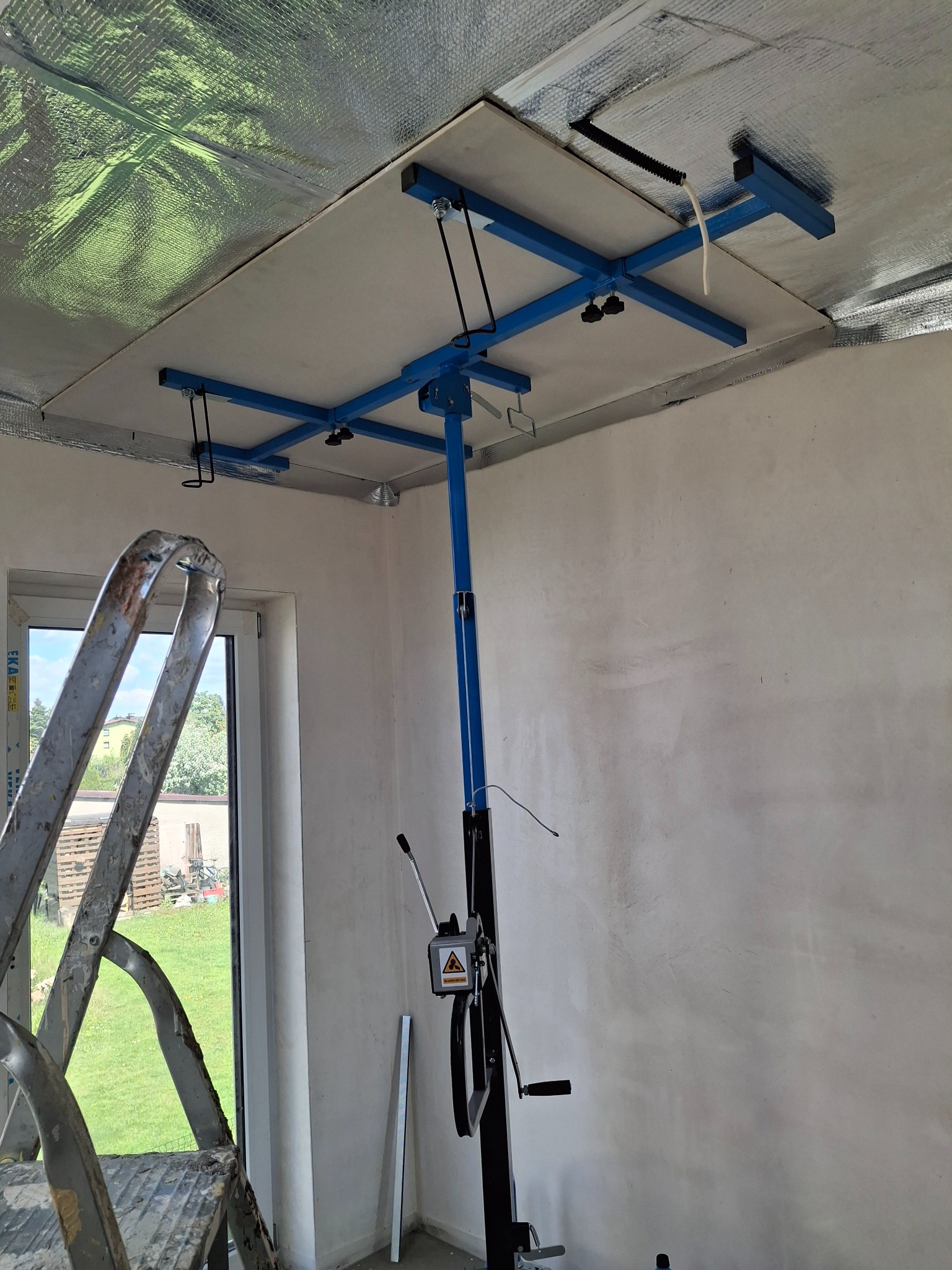 Montaż płyty karton gips na suficie z użyciem podnośnika. Widoczna drabina, okno i sufit z izolacją. Ujęcie z dołu, podkreślające etap prac.