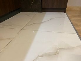 Błyszcząca, biała podłoga wyłożona płytkami imitującymi marmur z delikatnymi szarymi żyłkami, widoczne fragmenty mebli w tle.