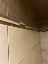 Ściana wyłożona jasnymi, kwadratowymi płytkami ceramicznymi z delikatną strukturą, u góry dekoracyjny pasek z mozaiki w odcieniach brązu i beżu, widoczny fragment sufitu z brązową listwą...