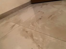 Błyszcząca, jasna podłoga z płytek imitujących marmur, zabrudzona resztkami zaprawy lub kleju po remoncie. Widoczne fugi i fragment przypodłogowej listwy.