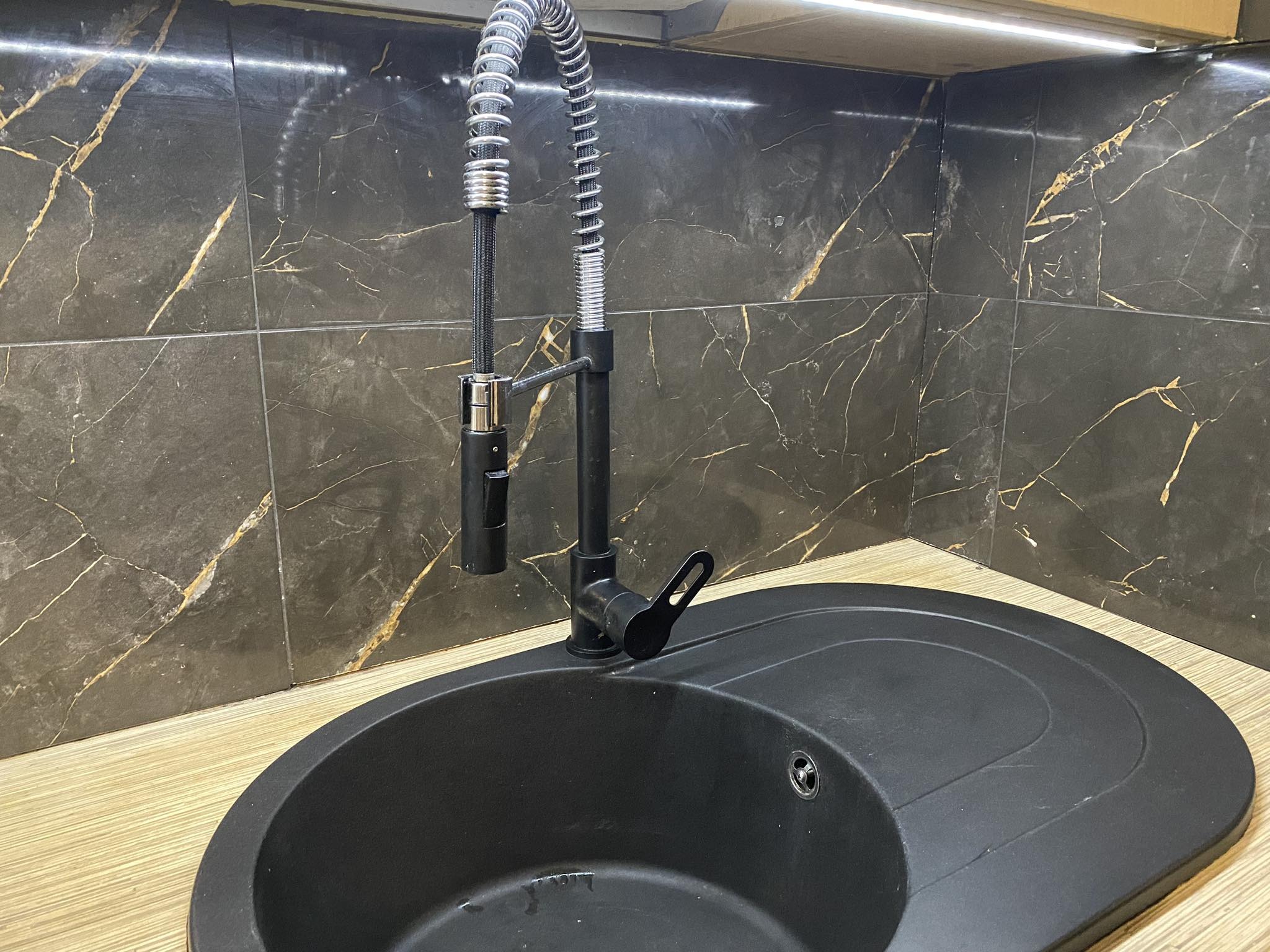 Czarny, okrągły zlew z baterią z wyciąganą wylewką na tle ściany wyłożonej ciemnymi płytkami z marmurkowym wzorem, oświetlonej od góry.