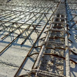 Zbrojenie fundamentu z widoczną siatką stalową i podkładkami dystansowymi. Jasne, kontrastowe oświetlenie podkreśla teksturę betonu i rdzawy odcień stali.