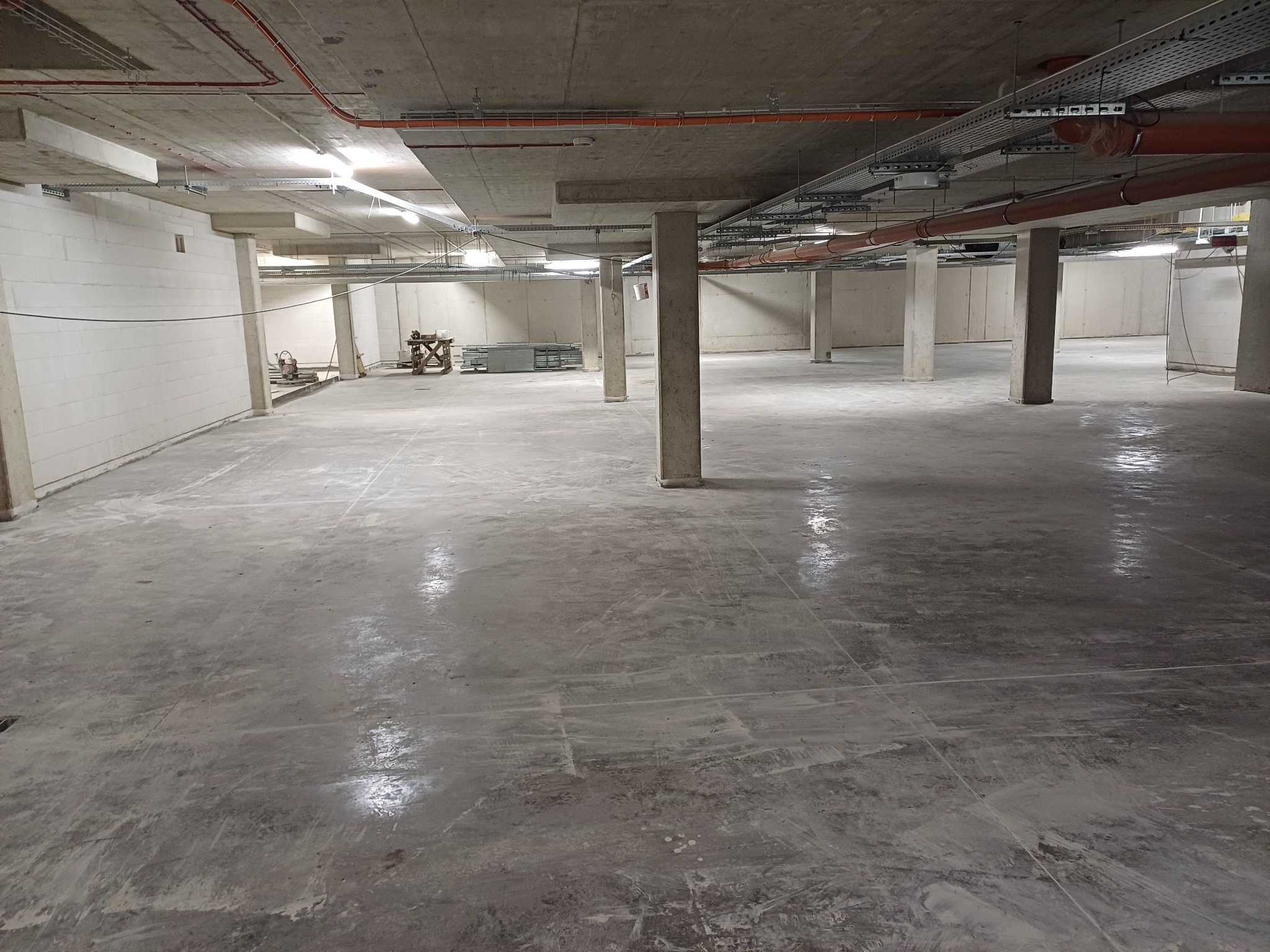 Surowy stan hali: betonowa podłoga, kolumny nośne, instalacje pod sufitem. Widoczne ślady budowy i przygotowania do dalszych prac wykończeniowych, surowy beton.