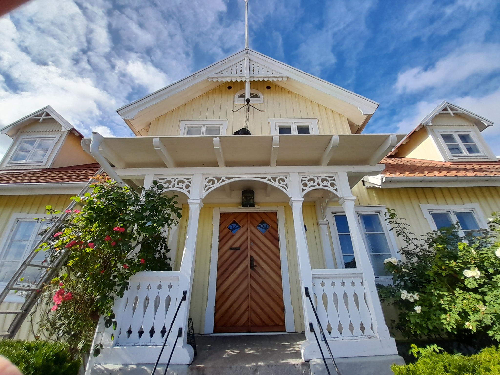 Żółty, dwupiętrowy dom z drewnianą fasadą i ozdobnymi detalami, widziany z dołu, z drabiną opartą o ścianę i kwitnącymi krzewami po bokach wejścia.