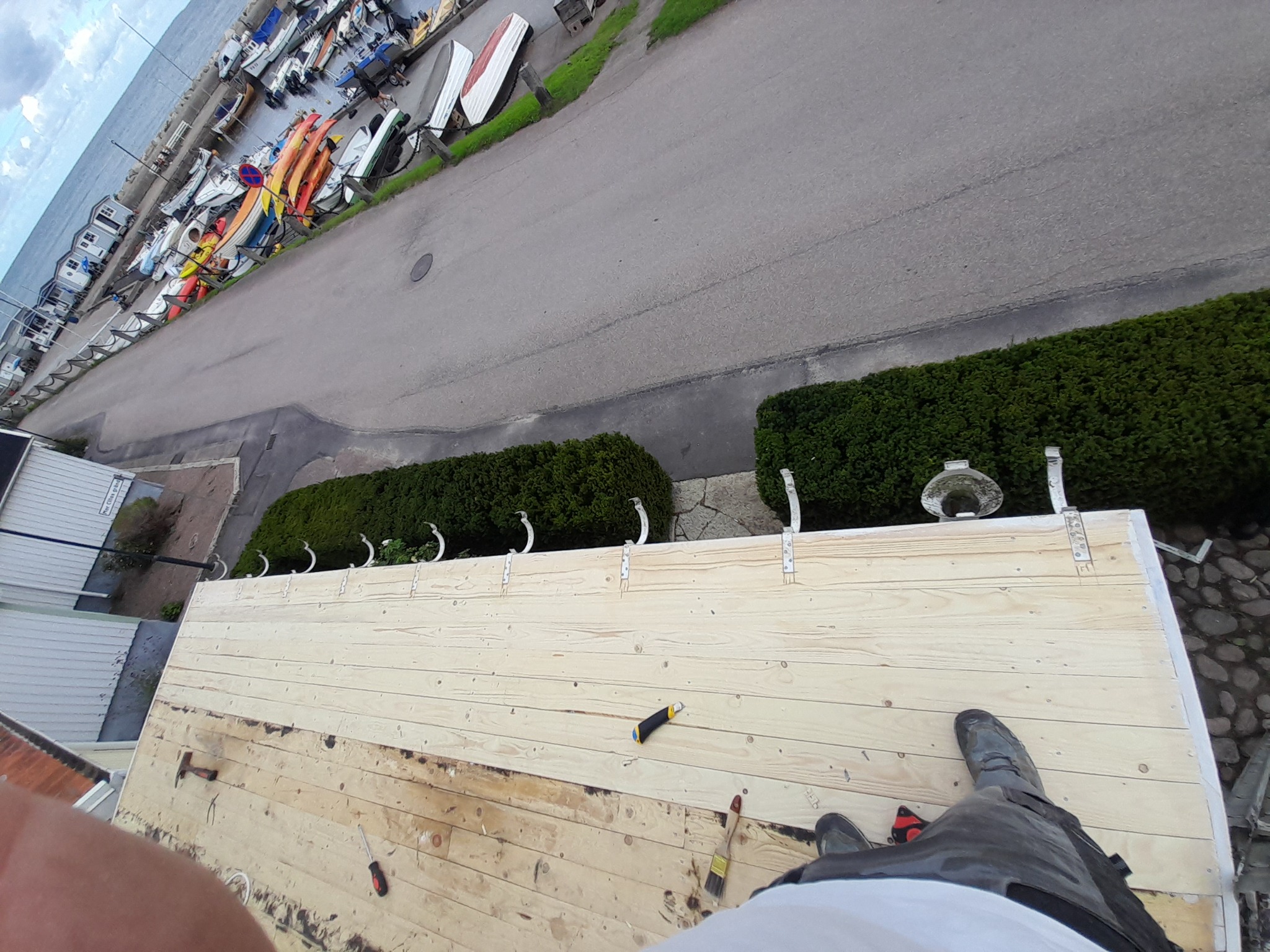 Widok z góry na drewnianą konstrukcję z widocznymi narzędziami (nóż, pędzel, śrubokręt) i fragmentem buta osoby pracującej, w tle port jachtowy i morze.