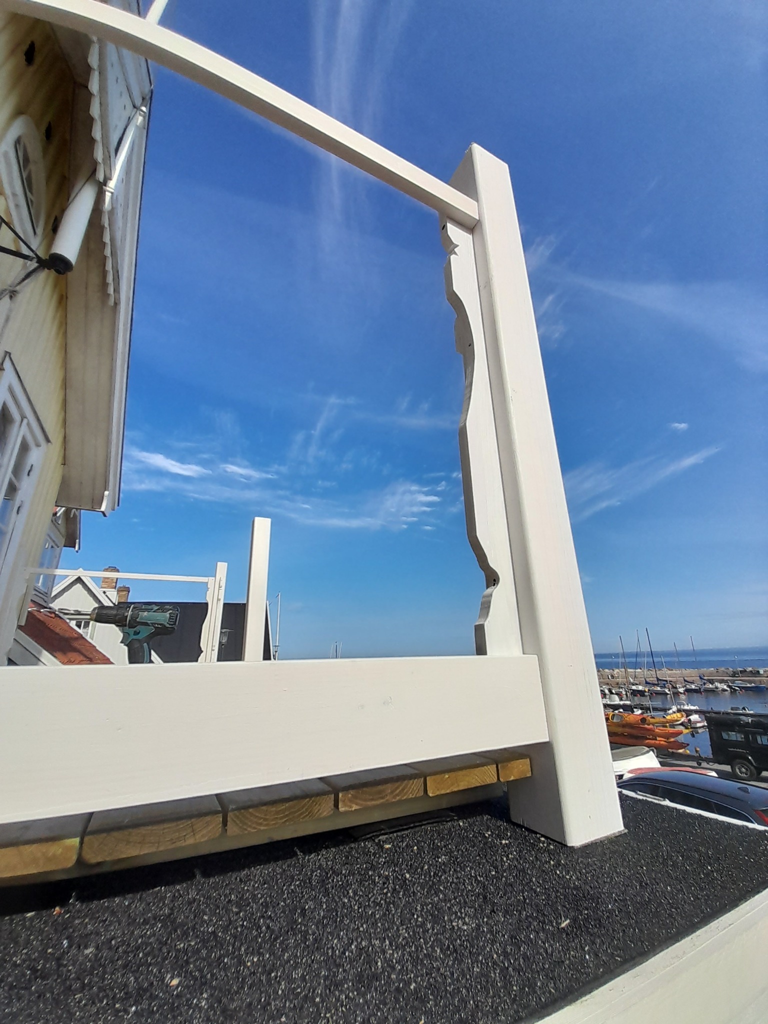 Montaż białej, drewnianej konstrukcji na dachu budynku, widok na port z łodziami i błękitne niebo. W tle widoczny wkrętak akumulatorowy.