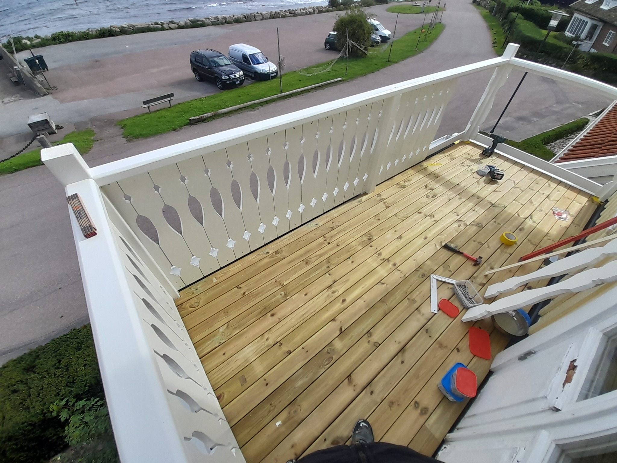 Nowy drewniany taras z białymi balustradami w trakcie budowy, widok z góry na narzędzia, farby i elementy konstrukcyjne, w tle widoczne budynki i drzewa.