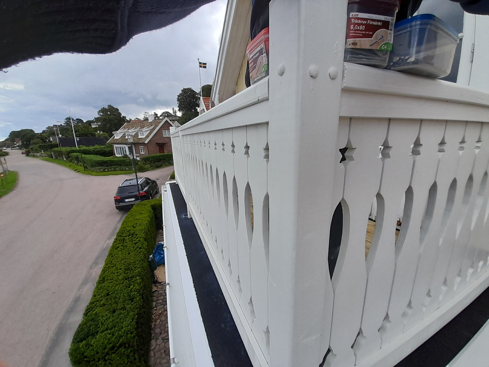Świeżo pomalowane, białe ogrodzenie drewniane z ozdobnymi wycięciami, widok z góry, na tle ulicy z samochodem i domami, pojemniki ze śrubami na górnej belce.