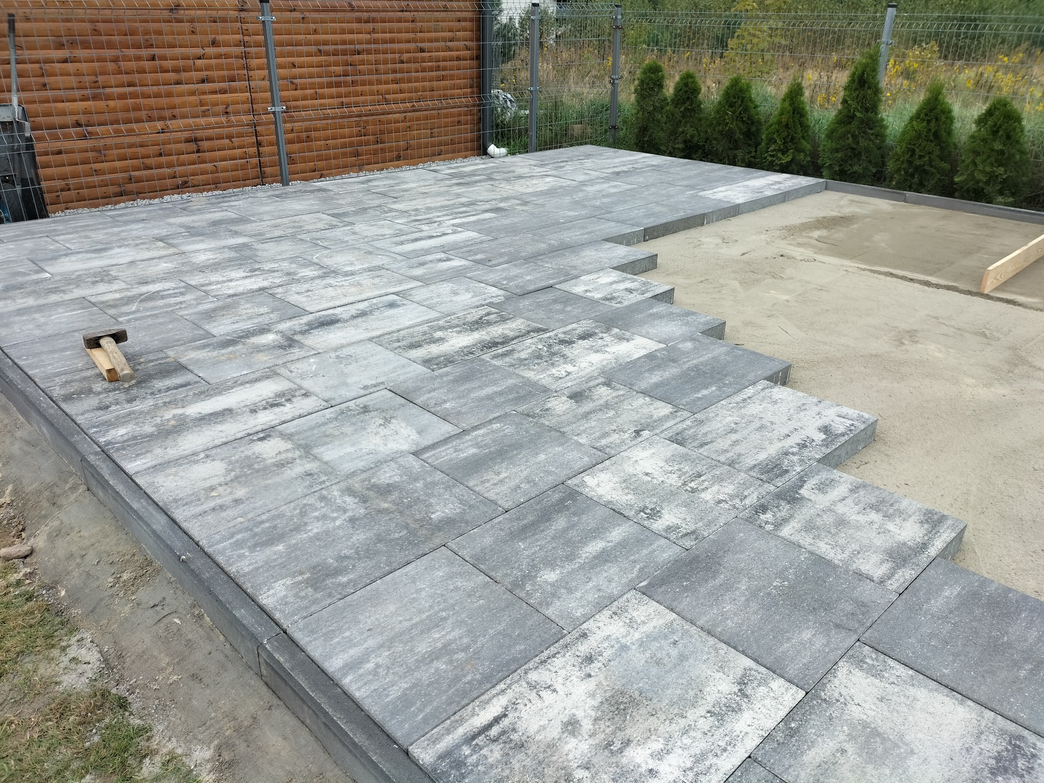 Szara kostka brukowa ułożona na tarasie, z widocznym młotkiem brukarskim, częściowo wykończona, z ogrodzeniem panelowym w tle i tujami.