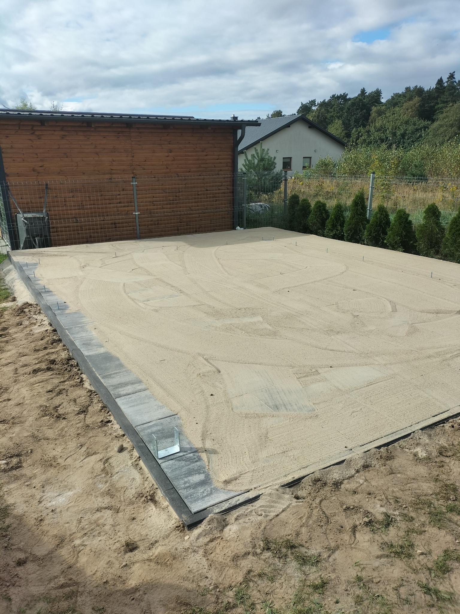 Przygotowany teren pod budowę tarasu: wyrównany piasek otoczony obrzeżami z szarej kostki, widoczne szpilki stabilizujące, w tle drewniany budynek gospodarczy i dom.