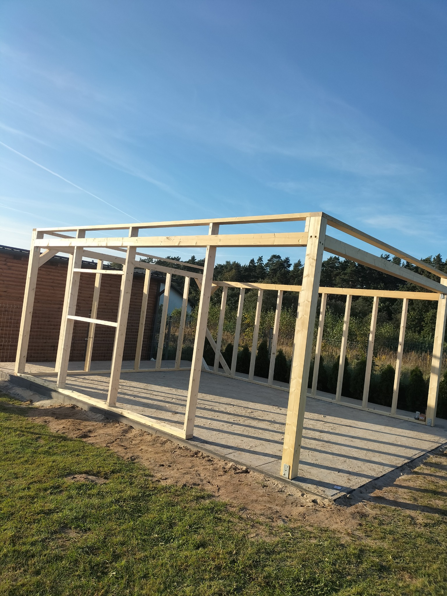 Szkielet drewnianej konstrukcji w trakcie budowy na betonowej płycie, widoczne elementy mocujące, w tle trawa, budynek i drzewa pod błękitnym niebem.