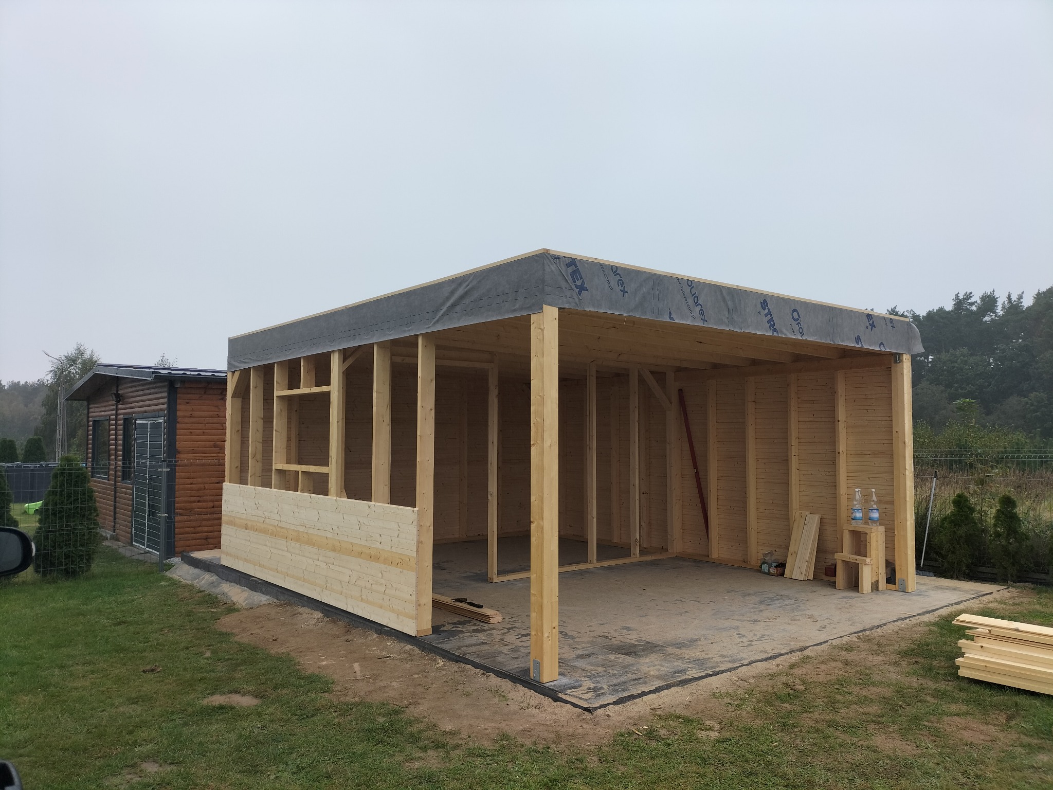 Budowa drewnianej konstrukcji z widocznym szkieletem, deski i izolacją dachową, obok gotowy budynek z drewna.