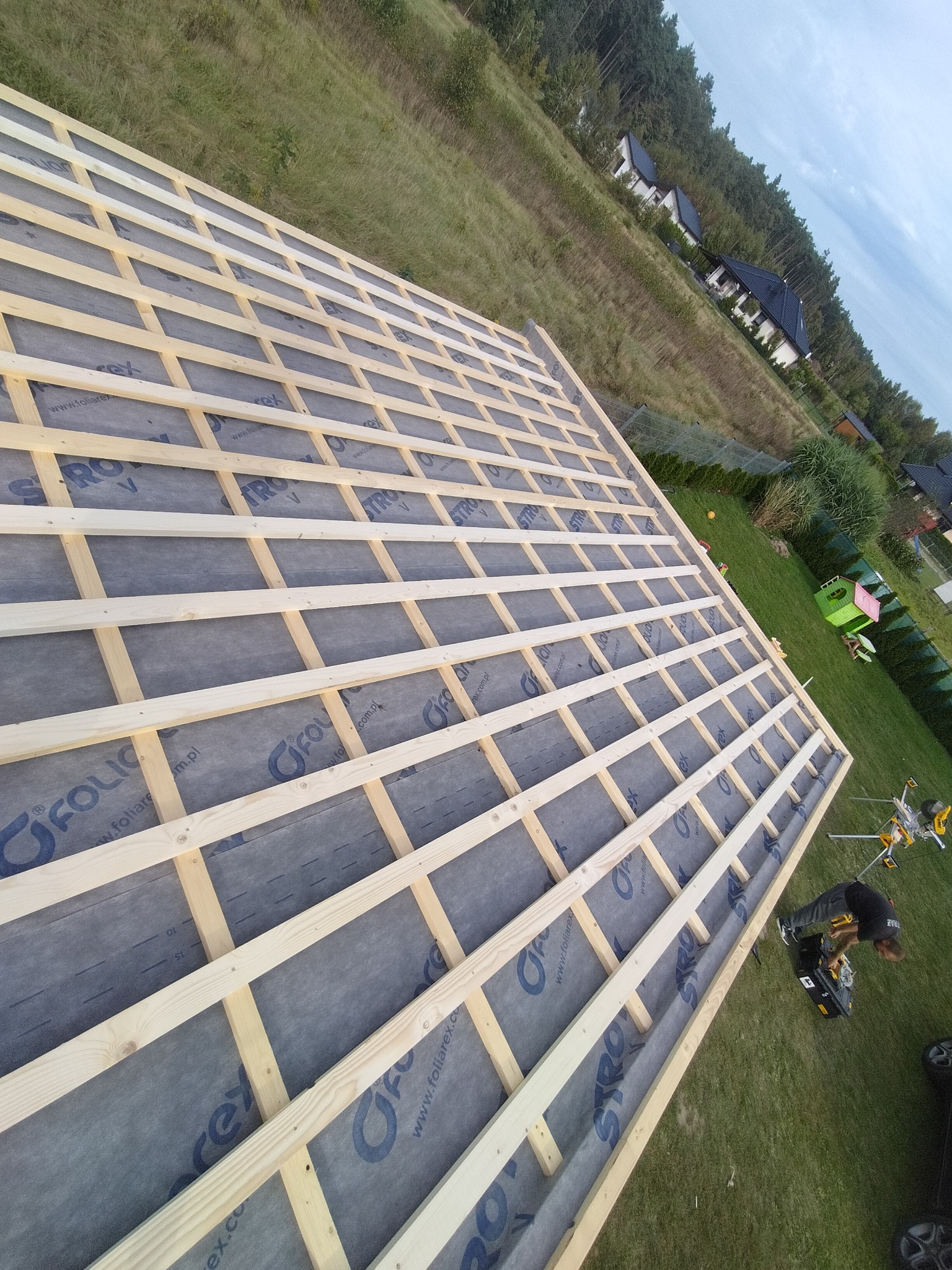 Konstrukcja dachu z drewnianych łat i kontrłat pokryta membraną dachową, widoczny pracownik z narzędziami, w tle domy i zieleń.