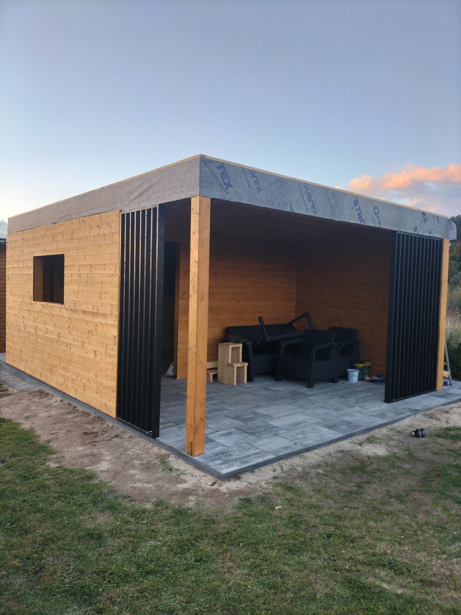 Nowoczesna drewniana altana w trakcie budowy, z czarnymi, pionowymi elementami dekoracyjnymi i przygotowanym miejscem do siedzenia wewnątrz, widoczne narzędzia i materiały budowlane.