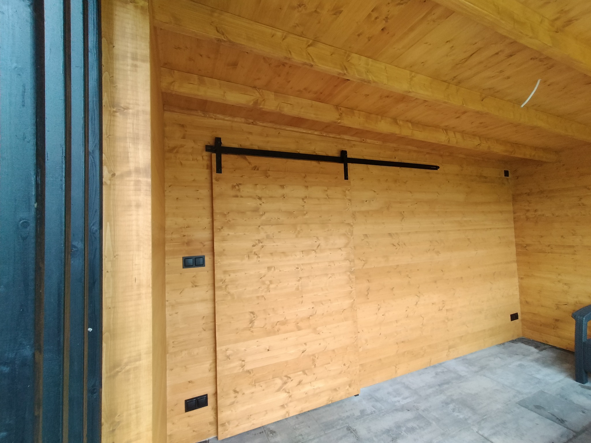 Drewniane panele ścienne i sufitowe w jasnym odcieniu z przesuwnymi drzwiami na czarnej szynie, widoczne gniazdka elektryczne i szare płytki podłogowe.